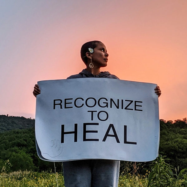 Artist Gina Goico spreads her arms holding a poster that reads, "Recognize to heal", in front of a hill against a pastel-coloured sky.